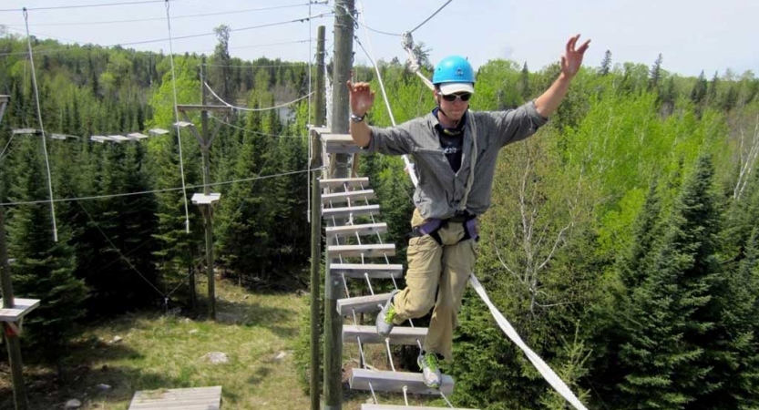 A person wearing safety gear and secured by ropes balances on an obstacle on a high ropes course. 
